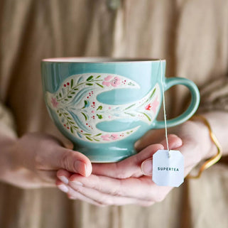 Person holding a decorative mug with a floral design, filled with hot water and a tea bag.