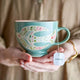 Person holding a decorative mug with a floral design, filled with hot water and a tea bag.