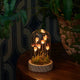 A decorative glass cloche dome containing glowing mushroom-like lights over moss and small stems, displayed on a wooden table beside a potted plant in a dim interior setting.
