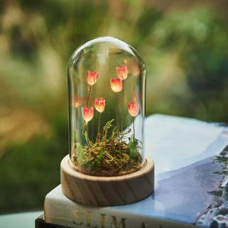 A small glass dome terrarium with glowing pink tulips and green moss sits on a round wooden base, displayed on top of a book outdoors with a blurred garden background.
