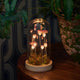 A decorative glass cloche dome lamp containing glowing pink mushrooms on a white base, placed on a wooden table next to a potted green plant in a dimly lit interior.