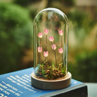 A small glass cloche dome with pink flowers and green moss sits on top of stacked books, with a softly blurred garden background.