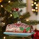 A holiday ornament depicting a chocolate roll with a Christmas tree on top, surrounded by glitter and red accents.