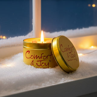 A gold-colored metal candle with a red wick, labeled 'Comfort & Joy' and 'Warm hearted wishes', is shown with its lid open.