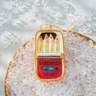 A decorative ornament shaped like an open sardine tin, with painted sardines above and the words 'SARDINES IN OIL' on a red front panel, displayed on a bed of clear decorative stones over a gold tray.