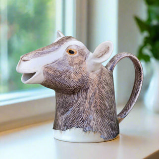 Goat-shaped jug on a wooden surface with greenery and flowers in the background