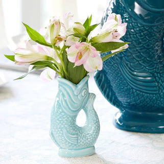 Teal fish-shaped vase on a table with flowers and fruit in the background