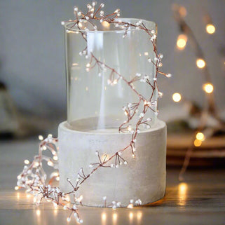 A glass vase with a decorative fairy light that consists of a cluster of white jewels surrounded by a warm white LED light, placed on a beige surface.