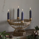A woven metal candle holder with five arms, holding lit dark blue taper candles, set on a rustic wooden surface with pine branches and pinecones against a textured plaster wall.