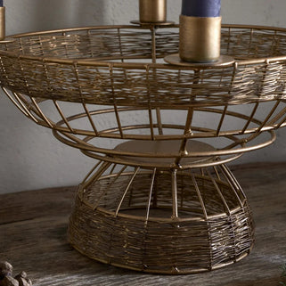 A brass-finished metal candelabra with a woven basket design, featuring a tiered structure and four candle holders, shown on a rustic wooden surface with pinecones and greenery.