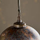 Close-up of a hanging pendant light with a spherical globe shade in distressed copper and brown tones, topped with an ornate brass cap and a dark cord against a softly blurred beige background.