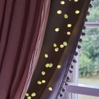 Warm white curtain string lights draped along the edge of a window, casting a soft glow against a dark curtain with pom-pom trim and a mauve curtain panel.