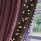 Warm white curtain string lights draped along the edge of a window, casting a soft glow against a dark curtain with pom-pom trim and a mauve curtain panel.