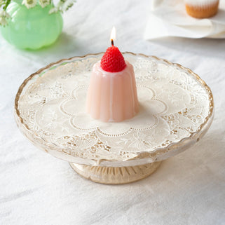 Pink candle with a red wick on a lace doily, with a vase of flowers and a cupcake in the background.