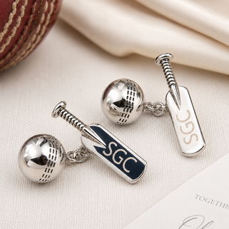 A pair of silver-toned cricket-themed cufflinks featuring a ball and a bat-shaped tag engraved with 'SGC', displayed on a textured cream fabric background with a red cricket ball and a card partially visible.