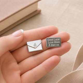 Two small metal charms, one shaped like an envelope and the other with text, held in a hand against a beige background.