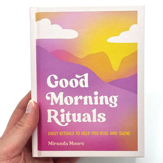 Book titled 'Good Morning Rituals' by Miranda Moore held by a hand with a colorful mountain design on the cover.