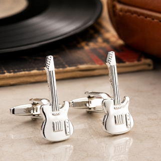 Silver guitar-shaped cufflinks on a reflective surface with a vinyl record and leather item in the background.