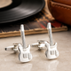 Silver guitar-shaped cufflinks on a reflective surface with a vinyl record and leather item in the background.