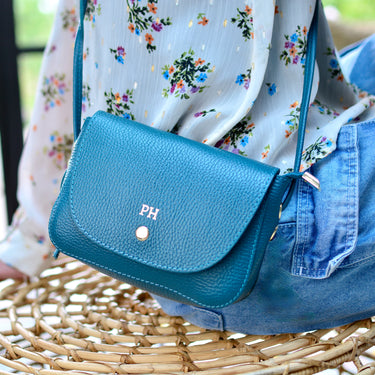 Teal leather saddle bag shown close up on model with gold foil monogram initials