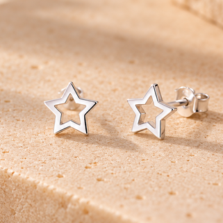 A pair of small silver star-shaped stud earrings resting on a textured beige stone surface with warm sunlight and shadows.