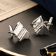 Silver cufflinks on a dark surface with a pen and paper in the background