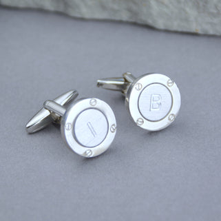 Silver porthole cufflinks with initial "I" on the left cufflink and "B" on the right cufflink, shown in close up shot on grey background.