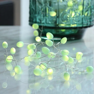 Green LED berry string lights draped over a translucent green glass vase on a reflective tabletop.
