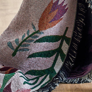 A close-up of a woven textile with a floral pattern, draped over a dark plush fabric with fringe trim, resting on a light wood floor.