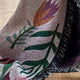 A close-up of a woven textile with a floral pattern, draped over a dark plush fabric with fringe trim, resting on a light wood floor.