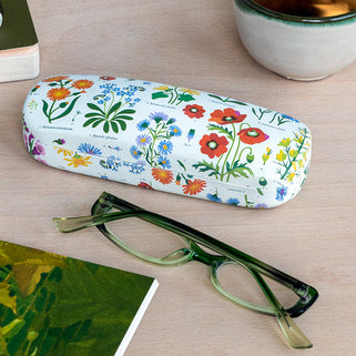 Floral-patterned glasses case with green glasses on a wooden surface with a mug of tea and book.
