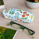Floral-patterned glasses case with green glasses on a wooden surface with a mug of tea and book.