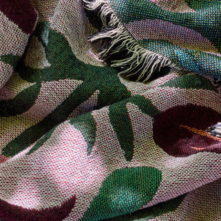 A close-up of a woven textile with a botanical pattern in green, burgundy, and lavender on a light pink base, featuring fringed edges.