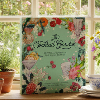 Book titled 'The Cocktail Garden' on a windowsill with potted plants and flowers.