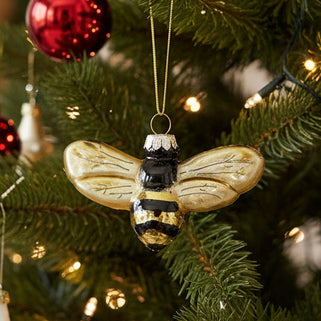 A golden glass bauble in the shape of a bee with gold and black details, hanging from a tree.