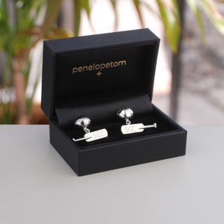 Cricket bat and ball cufflinks with personalised "SGC" initials engraved on both bats, shown in black Penelopetom cufflinks box.