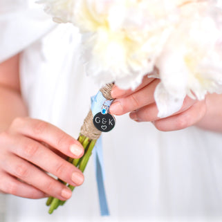 Gunmetal round wedding bouquet keepsake charm with personalised message "G&K heart"  engraved on the front and Blue Topaz/September birthstone. Shown with blue ribbon wrapped around bridal flowers.