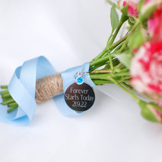 Gunmetal round wedding bouquet keepsake charm with personalised message "Forever Starts Today 21.9.22"  engraved on the front and Blue Topaz/September birthstone. Shown with blue ribbon wrapped around bridal flowers.