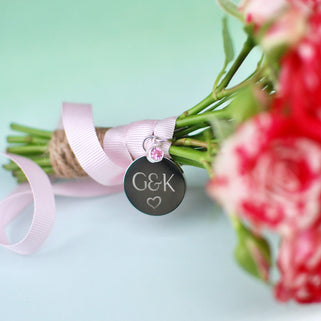 Gunmetal round wedding bouquet keepsake charm with personalised message "G&K heart"  engraved on the front and Alexandrite/June birthstone. Shown with pink ribbon wrapped around bridal flowers.