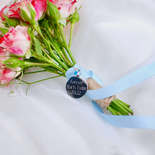 Silver round wedding bouquet keepsake charm with personalised message "Forever Starts Today 21.9.22"  engraved on the front and blue topaz/December birthstone. Shown with blue ribbon wrapped around bridal flowers.
