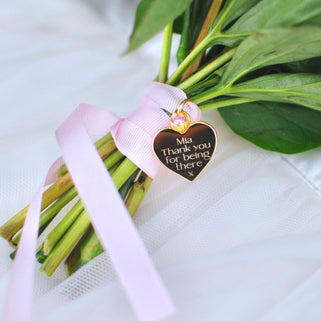 Gold heart wedding bouquet keepsake charm with personalised message "Mia Thank you for being there x"  engraved on the front and Alexandrite/June birthstone. Shown with pink ribbon wrapped around bridal flowers.