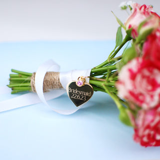 Gold heart wedding bouquet keepsake charm with personalised message "Bridesmaid 22.6.23"  engraved on the front and Alexandrite/June birthstone. Shown with white ribbon wrapped around bridal flowers.