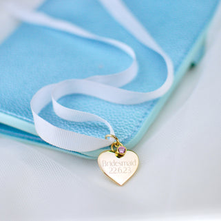 Gold heart wedding bouquet keepsake charm with personalised message "Bridesmaid 22.6.23"  engraved on the front and Alexandrite/June birthstone. Shown with white ribbon in close up shot