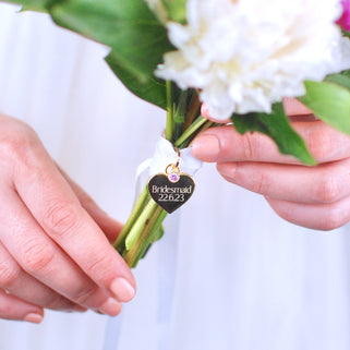 Gold heart wedding bouquet keepsake charm with personalised message "Bridesmaid 22.6.23"  engraved on the front and Alexandrite/June birthstone. Shown with white ribbon wrapped around bridal flowers.