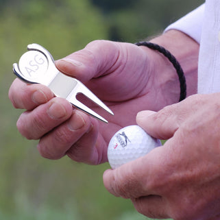 Golf divet repair and ball marker tool marker with "ASG" initials engraved, shown in close up shot with model holding golf ball.