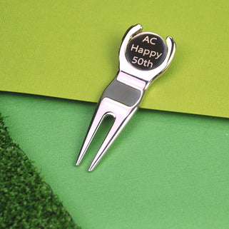 Golf divet repair and ball marker tool with "AC Happy 50th" engraved, shown in close up shot on green background.