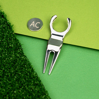 Golf divet repair and ball marker tool with "AC" engraved, shown in close up shot on green background.