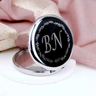 Silver compact mirror with personalised "BN" initials engraved in the centre with a floral wreath border, shown in close up shot with pink background