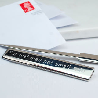 Silver letter opener with "For real mail not email heart Jo" engraved on  the front, shown in close up shot with a letter.