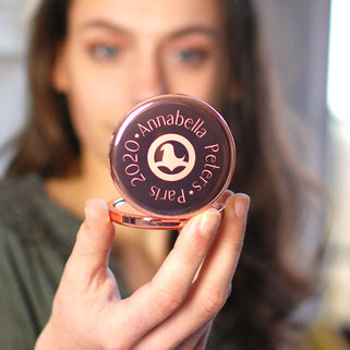Rose gold compact mirror with personalised logo in the centre and text engraved around the outside, shown in close up with model.